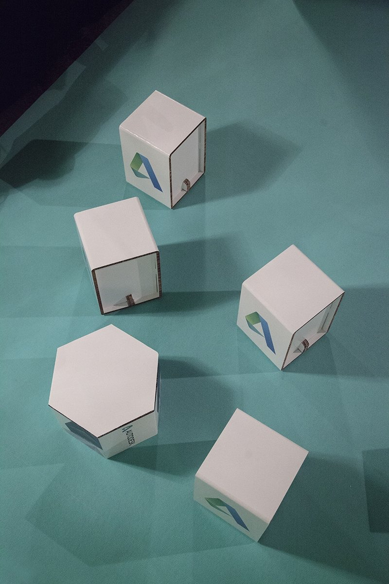 Cinco taburetes de cartón blanco con forma cúbica y hexagonal sobre fondo verde, usados como mobiliario ecológico en eventos sostenibles