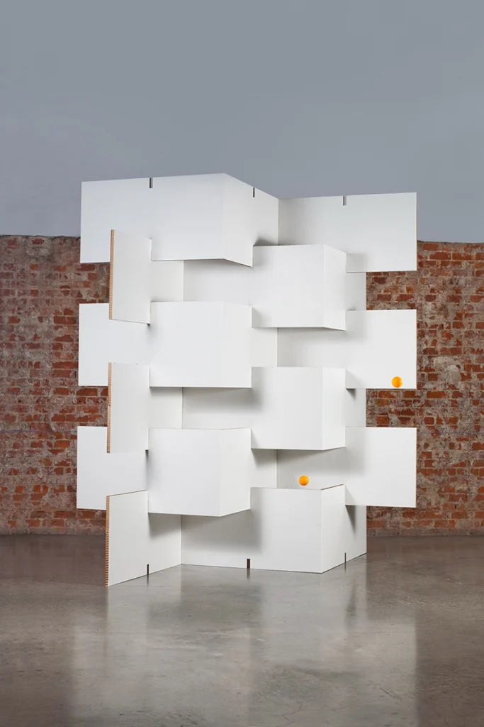 Biombo de cartón blanco BYÓ con diseño modular en zigzag colocado frente a una pared de ladrillo visto sobre suelo de cemento pulido