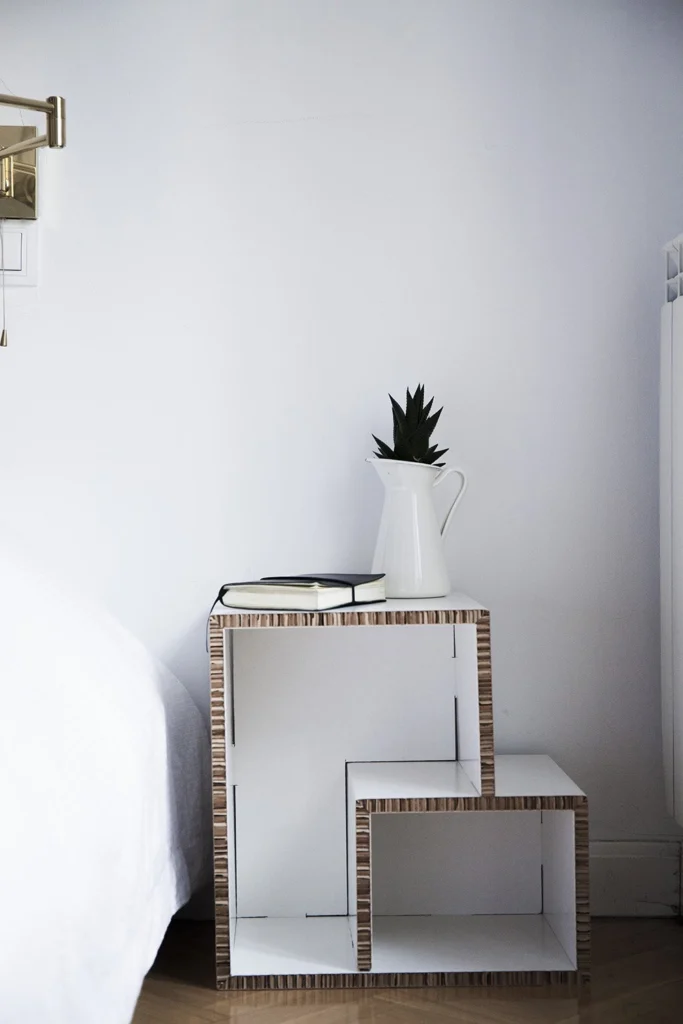 Estantería modular de cartón usada como mesita de noche junto a una cama, estilo vintage, con planta y cuaderno encima