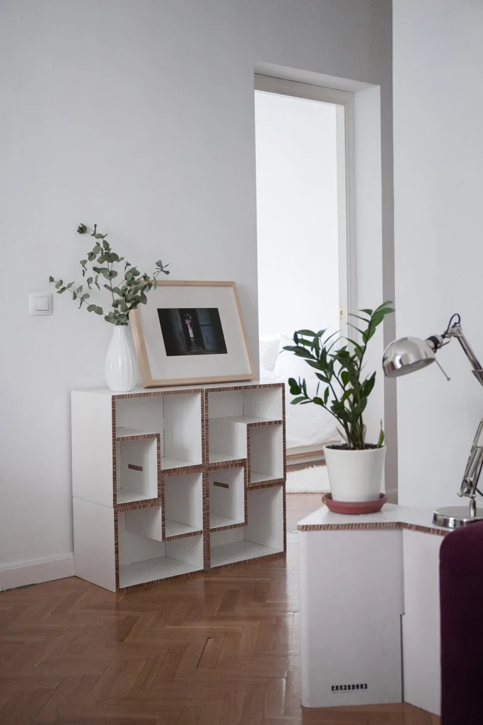 Estantería modular de cartón usada como mesita de noche junto a una cama, estilo vintage, con planta y cuaderno encima