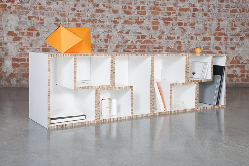 Estantería modular de cartón sobre suelo de hormigón con fondo de ladrillo visto y elementos decorativos coloridos